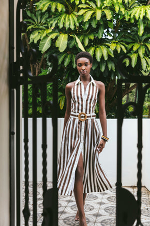 A woman wears the Cara Cara Quentin Midi Dress, featuring brown and white stripes and a belted waist, as she walks on patterned tile with green plants and a white wall in the background.