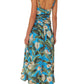 A person is wearing a Farm Rio Blue Anthurium Garden Maxi Dress made from eco-responsible fibers, featuring a blue botanical print with thin straps and an open back, paired with blue heels. The person is standing facing away from the camera.