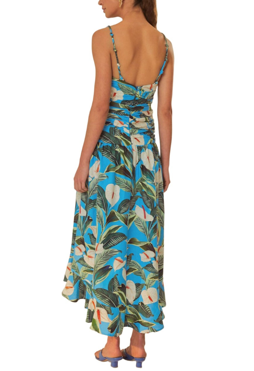 A person is wearing a Farm Rio Blue Anthurium Garden Maxi Dress made from eco-responsible fibers, featuring a blue botanical print with thin straps and an open back, paired with blue heels. The person is standing facing away from the camera.