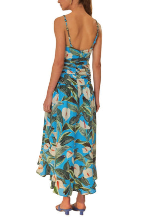 A person is wearing a Farm Rio Blue Anthurium Garden Maxi Dress made from eco-responsible fibers, featuring a blue botanical print with thin straps and an open back, paired with blue heels. The person is standing facing away from the camera.