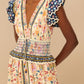 Model wears the Farm Rio Superbloom Tapestry Cream Maxi Dress with smocked waist and ruffled sleeves featuring a blue geometric pattern, styled with gold earrings, set against a beige background.