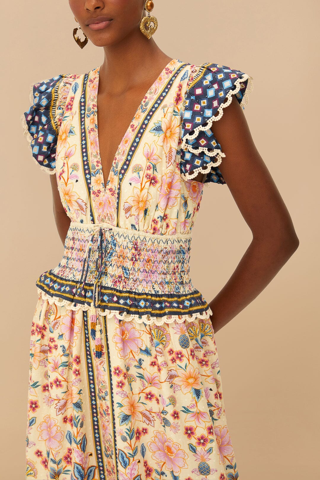 Model wears the Farm Rio Superbloom Tapestry Cream Maxi Dress with smocked waist and ruffled sleeves featuring a blue geometric pattern, styled with gold earrings, set against a beige background.