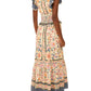 A woman is seen from behind wearing the Farm Rio Superbloom Tapestry Cream Maxi Dress with ruffled sleeves and hem, paired with yellow shoes and heart-shaped earrings.