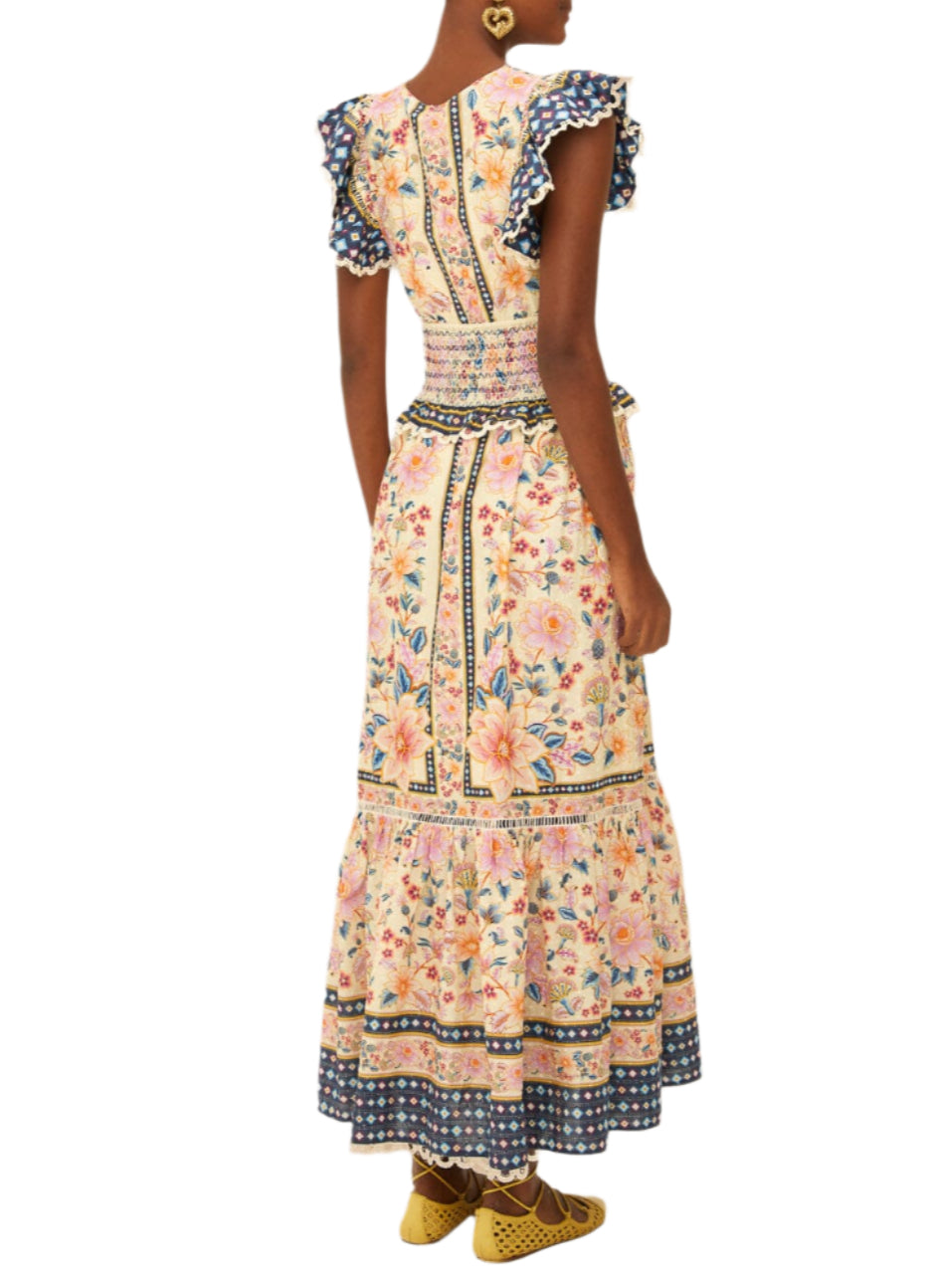 A woman is seen from behind wearing the Farm Rio Superbloom Tapestry Cream Maxi Dress with ruffled sleeves and hem, paired with yellow shoes and heart-shaped earrings.