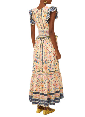 A woman is seen from behind wearing the Farm Rio Superbloom Tapestry Cream Maxi Dress with ruffled sleeves and hem, paired with yellow shoes and heart-shaped earrings.