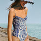 A woman models the Farm Rio FR Delicate Tapestry One Piece swimsuit and a striped sunhat while standing on a rocky shore with the ocean behind her.