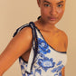 A woman with braided hair models the Farm Rio Blue Floral Cascade One Piece, a one-shoulder swimsuit with blue florals and dark blue crochet trim, exuding beach elegance against a beige background.