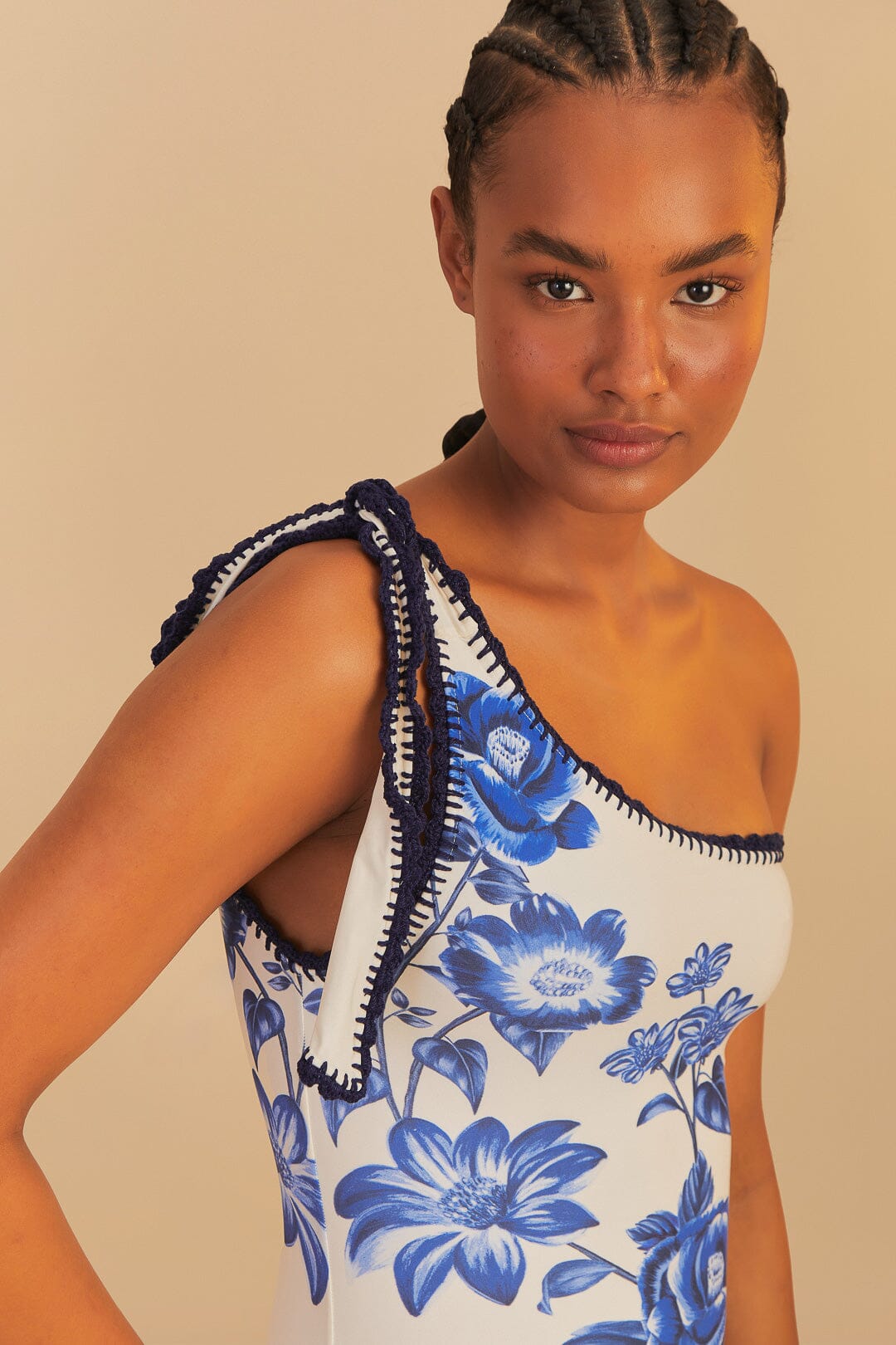 A woman with braided hair models the Farm Rio Blue Floral Cascade One Piece, a one-shoulder swimsuit with blue florals and dark blue crochet trim, exuding beach elegance against a beige background.