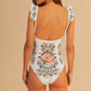 A woman models the Farm Rio Sweet Bloom Bouquet One Piece, an off-white swimsuit with ruffled straps and vibrant floral prints, shown from the back against a beige background.