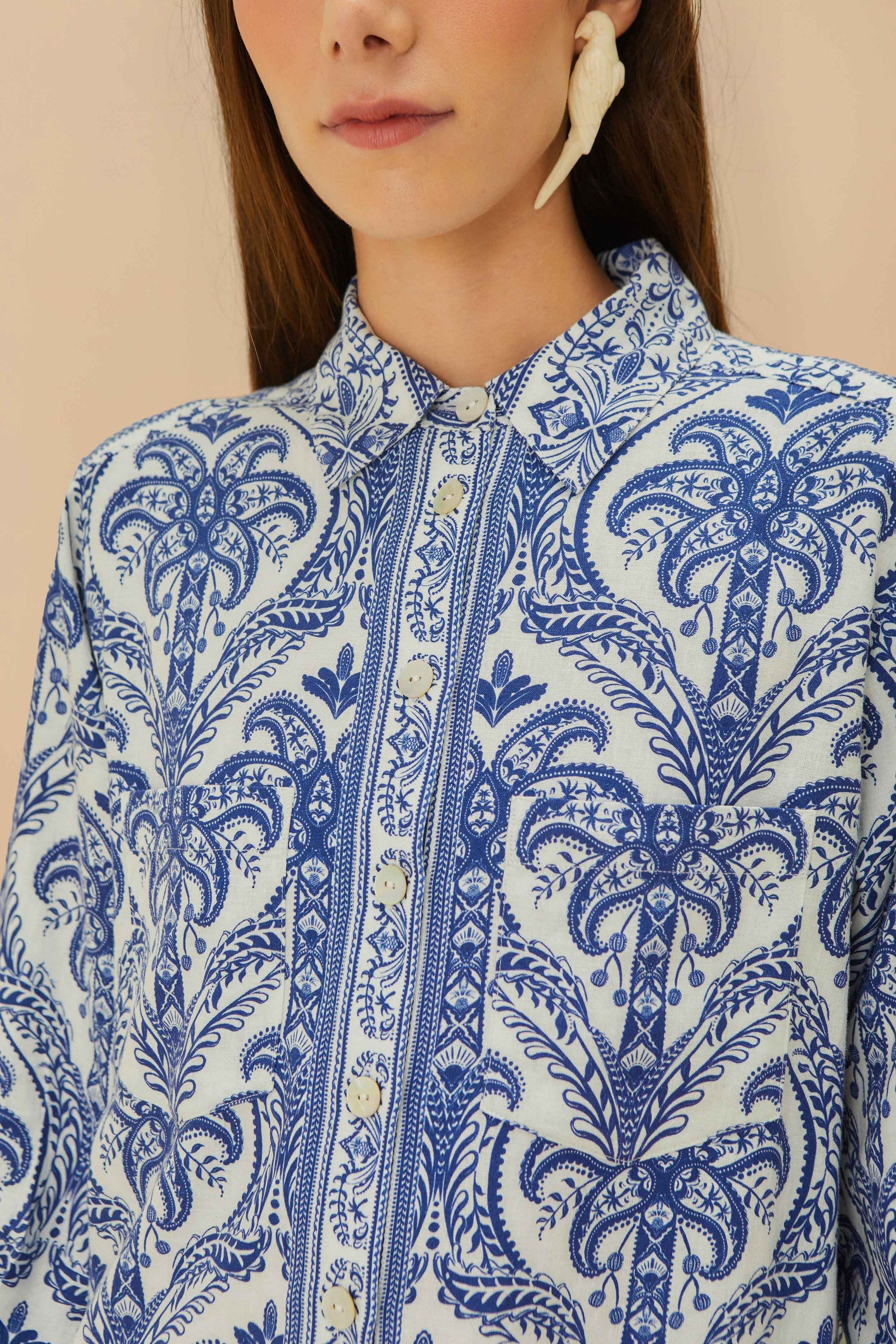 A woman wears the Farm Rio Blue Tropical Cameo Off-White Pajama Shirt, featuring an intricate floral pattern and eco-friendly LENZING™ ECOVERO™, paired with a large off-white earring.