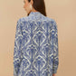 A woman with long brown hair, seen from behind, wears the Farm Rio Blue Tropical Cameo Off-White Pajama Shirt, featuring blue and white patterns, as she stands against a beige background.