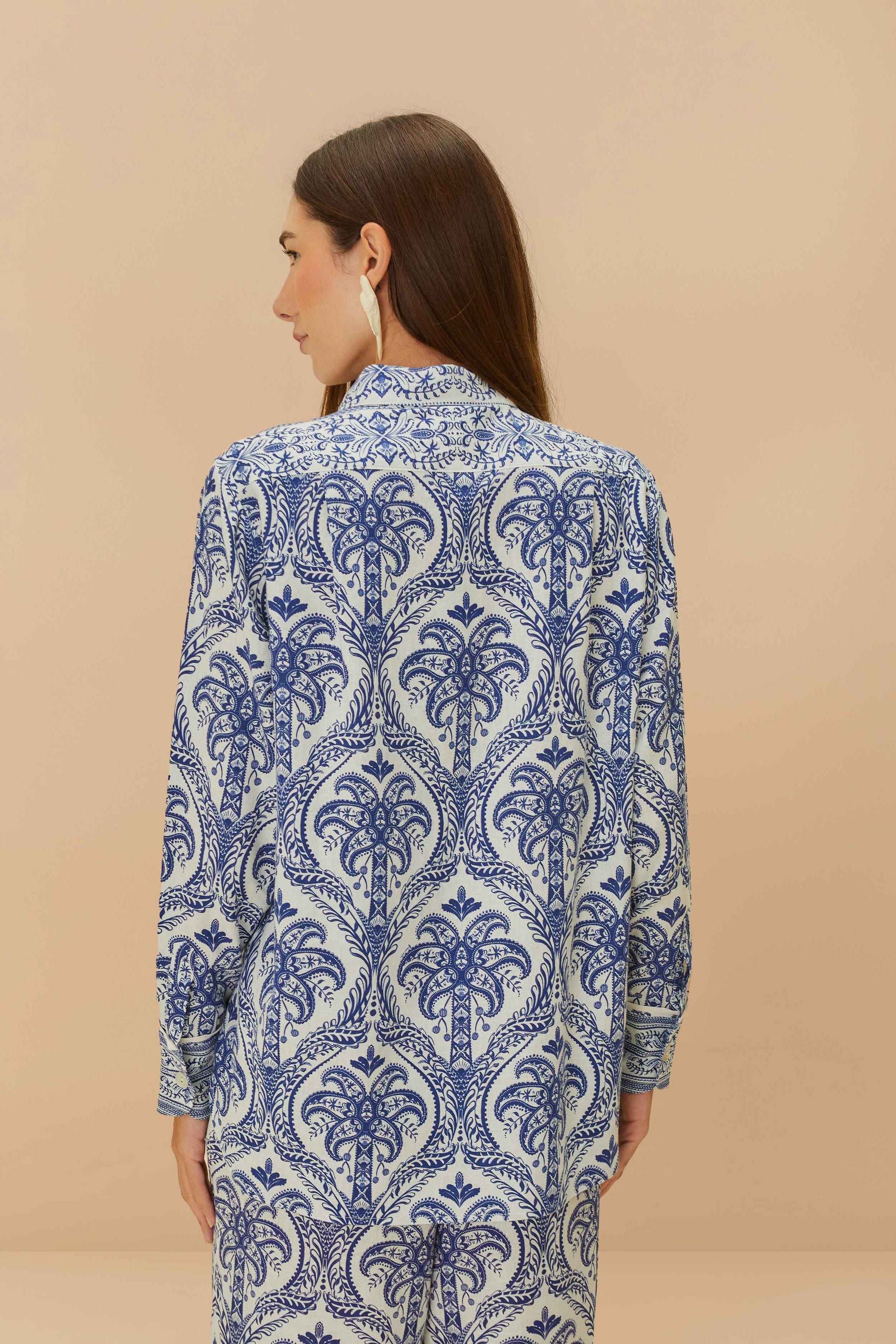 A woman with long brown hair, seen from behind, wears the Farm Rio Blue Tropical Cameo Off-White Pajama Shirt, featuring blue and white patterns, as she stands against a beige background.