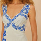 A woman wears the Farm Rio Off-White Floral Cascade Wavy Midi Dress, featuring blue floral and striped patterns on an off-white background, shown from shoulders to mid-thigh.