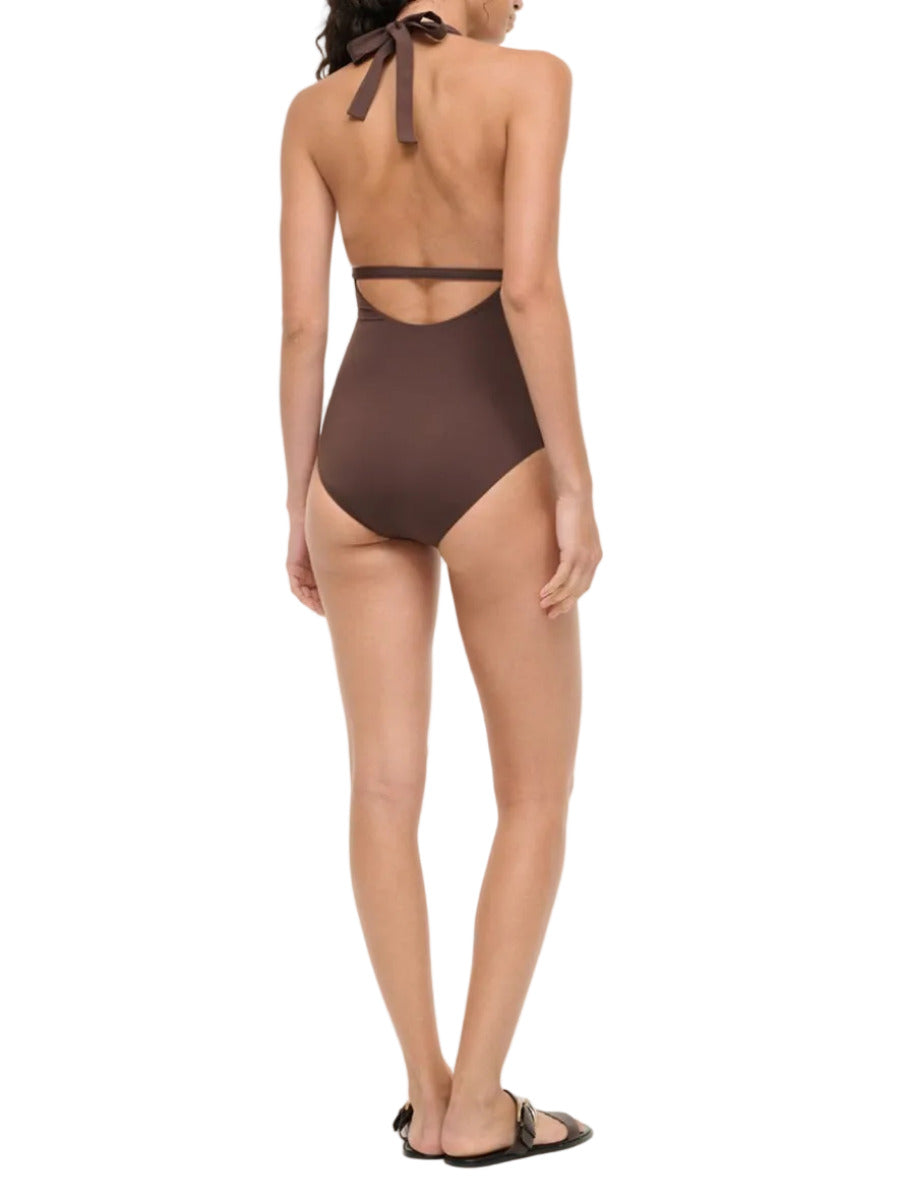 A woman wears the Staud La Jolla One Piece, a brown halter swimsuit with a moon buckle detail, open back, and horizontal back strap, paired with sandals.