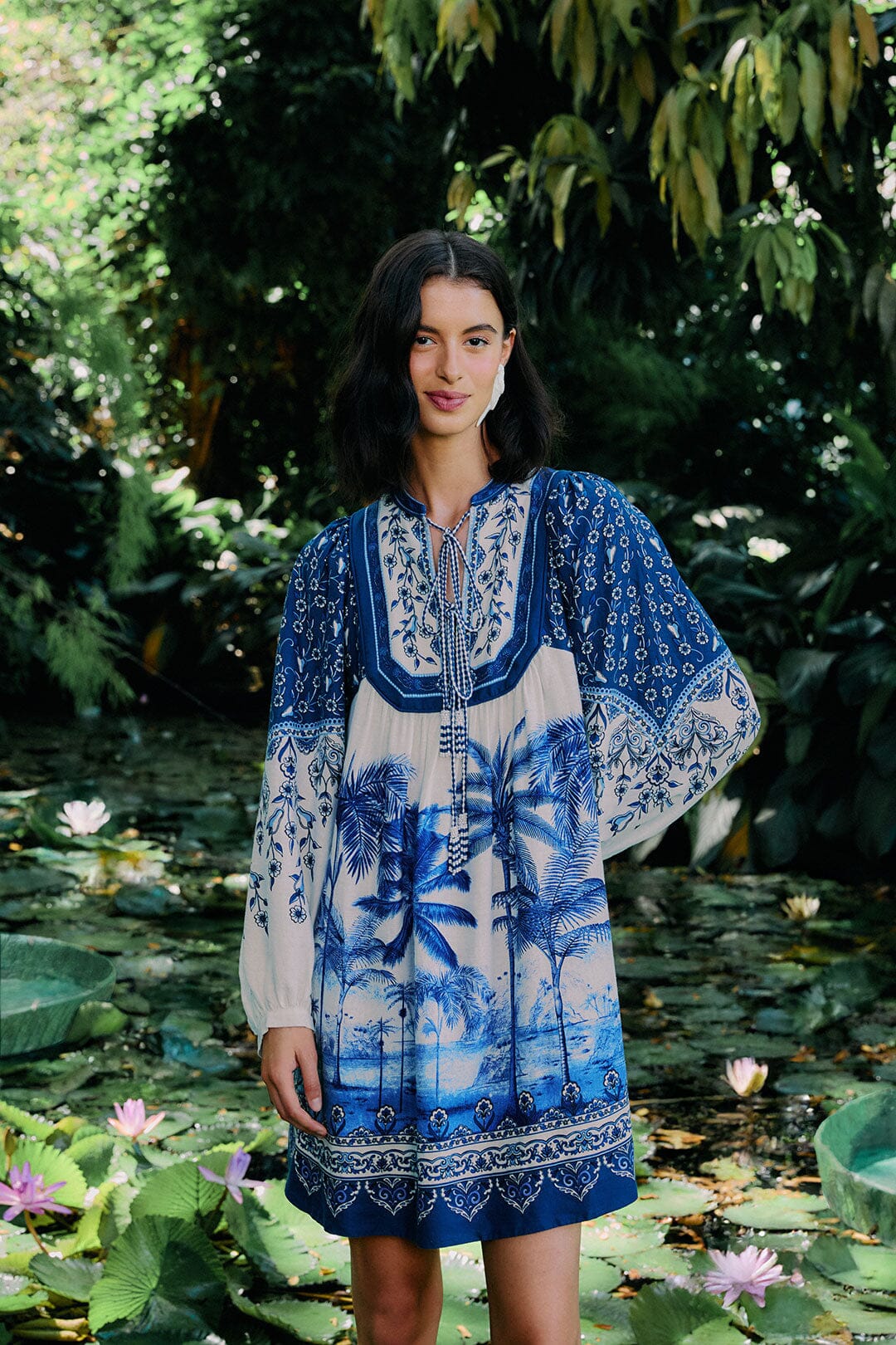 A woman stands by a pond with lily pads, wearing the Farm Rio Palm Tree Beach Off-White Mini Dress, featuring long sleeves and a vibrant tropical pattern that embodies lush, exotic style.