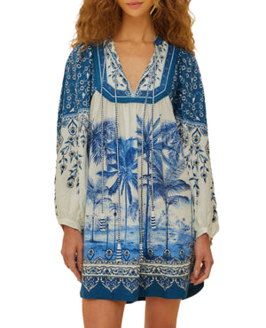 Woman wearing a Farm Rio Palm Tree Beach Off-White Mini Dress with a tropical palm tree and floral print, made from eco-responsible LENZING™ ECOVERO™ fibers, standing against a white background.
