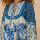 A woman wears a Farm Rio Palm Tree Beach Off-White Mini Dress featuring blue and white floral and palm tree designs, long sleeves, and a tie neckline—embodying tropical beauty in a chic style.