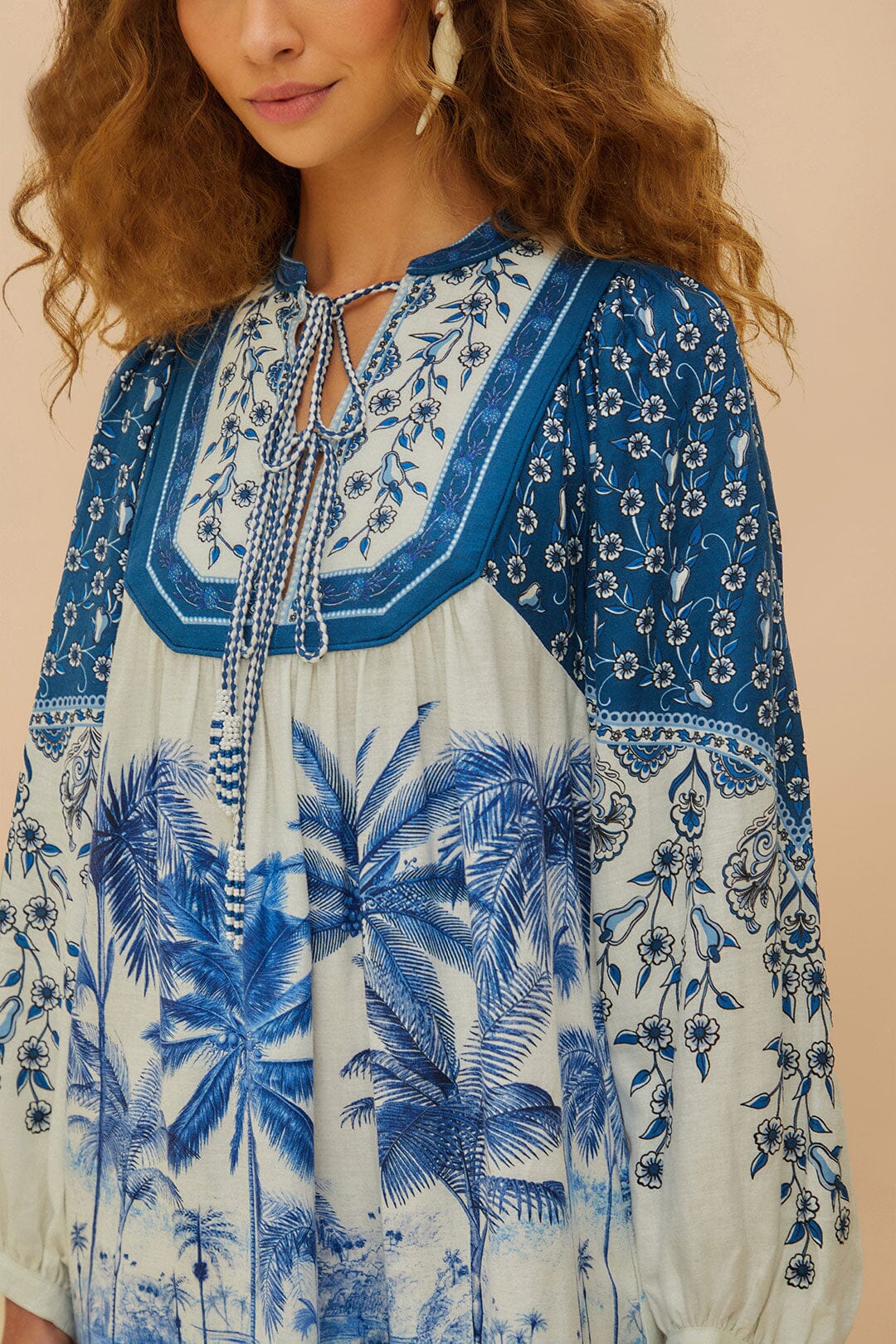 A woman wears a Farm Rio Palm Tree Beach Off-White Mini Dress featuring blue and white floral and palm tree designs, long sleeves, and a tie neckline—embodying tropical beauty in a chic style.
