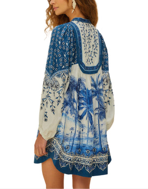 A woman showcases the Palm Tree Beach Off-White Mini Dress by Farm Rio—crafted from eco-responsible fibers, this loose blue and white long-sleeve piece features a vibrant palm tree print, radiating effortless tropical style from the back.