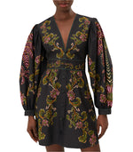 A person is wearing a stunning Farm Rio Ravenala Black V Neckline Mini Dress, with long sleeves featuring intricate green, pink, and gold floral patterns. This elegant look epitomizes FARM Rio's signature style.