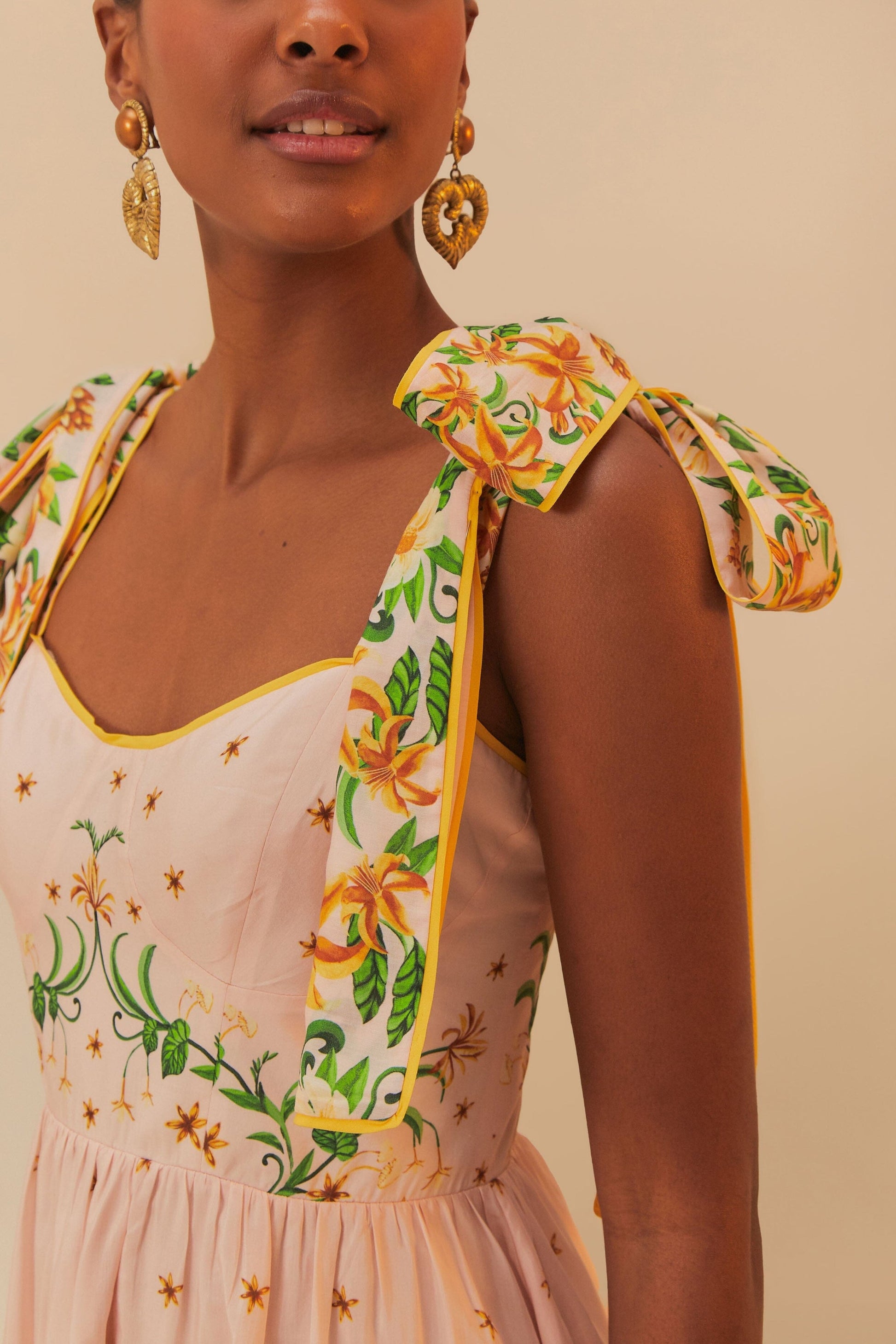 A woman in the Farm Rio Botanical Beauty Shoulder Bow Midi Dress, with botanical prints and shoulder bows, accessorizes with gold heart-shaped earrings.