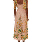 A woman faces away, wearing the Farm Rio Botanical Beauty Shoulder Bow Midi Dress in light pink with a vibrant botanical pattern. She pairs it with gold earrings and brown flat shoes.
