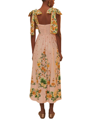 A woman faces away, wearing the Farm Rio Botanical Beauty Shoulder Bow Midi Dress in light pink with a vibrant botanical pattern. She pairs it with gold earrings and brown flat shoes.