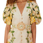 A woman wears the Farm Rio Garden Cascade Sand Mini Dress featuring a cream and yellow floral print, puffed sleeves, deep V-neckline, button front, and a matching belt with circular buckle.