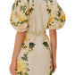 A woman is shown from the back wearing the Farm Rio Garden Cascade Sand Mini Dress, a short, light-colored dress with puffy sleeves and a yellow floral pattern.