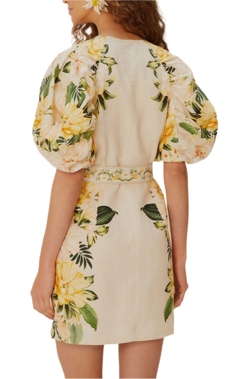 A woman is shown from the back wearing the Farm Rio Garden Cascade Sand Mini Dress, a short, light-colored dress with puffy sleeves and a yellow floral pattern.