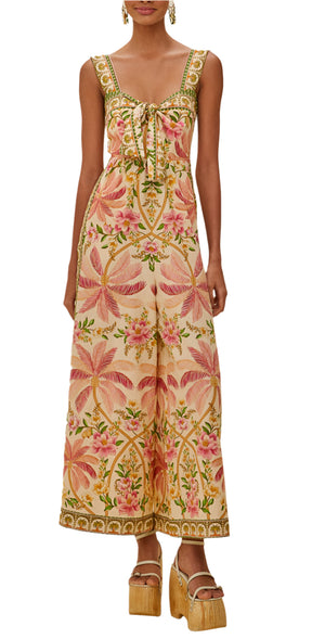 Woman in the Palm Dream Off White Jumpsuit by Farm Rio—sleeveless, long, with pink floral and palm print, front tie detail, and gold platform sandals—perfectly capturing laid-back tropical vibes.