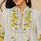 A woman wears a Farm Rio Sicily Scarf Off White Mini Dress, detailed with yellow floral embroidery, blue borders, and a tie neckline.