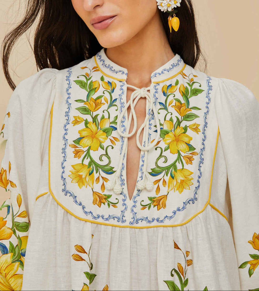 A woman wears a Farm Rio Sicily Scarf Off White Mini Dress, detailed with yellow floral embroidery, blue borders, and a tie neckline.