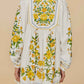 A woman wears the Sicily Scarf Off White Mini Dress by Farm Rio, a white embroidered summer dress with long sleeves and yellow-green floral details on the back, sleeves, and hem.