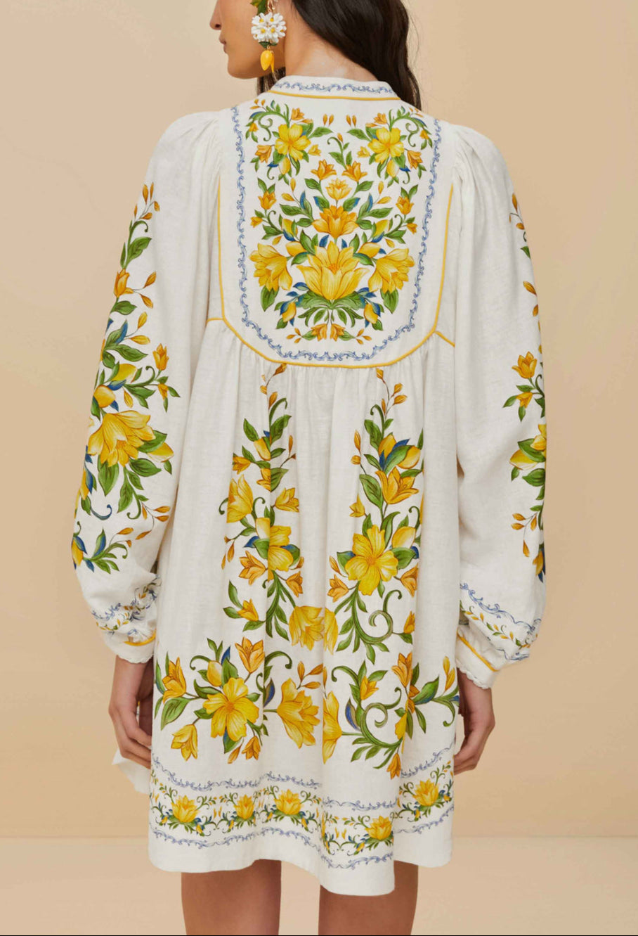 A woman wears the Sicily Scarf Off White Mini Dress by Farm Rio, a white embroidered summer dress with long sleeves and yellow-green floral details on the back, sleeves, and hem.