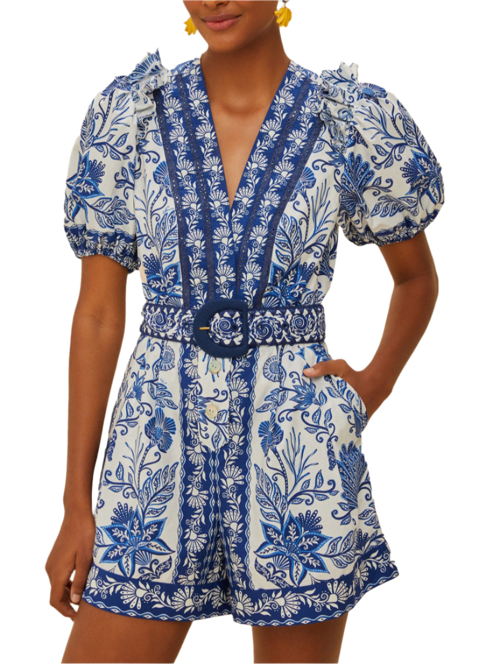 A person wears the Shell Garden Off White Romper by Farm Rio, featuring tropical florals, puffed short sleeves, a V-neckline, and a matching belted waist.