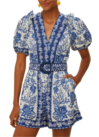 A person wears the Shell Garden Off White Romper by Farm Rio, featuring tropical florals, puffed short sleeves, a V-neckline, and a matching belted waist.