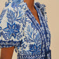 A woman wears the Farm Rio Shell Garden Off White Romper with blue florals, puff sleeves, a belted waist, and yellow floral earrings.