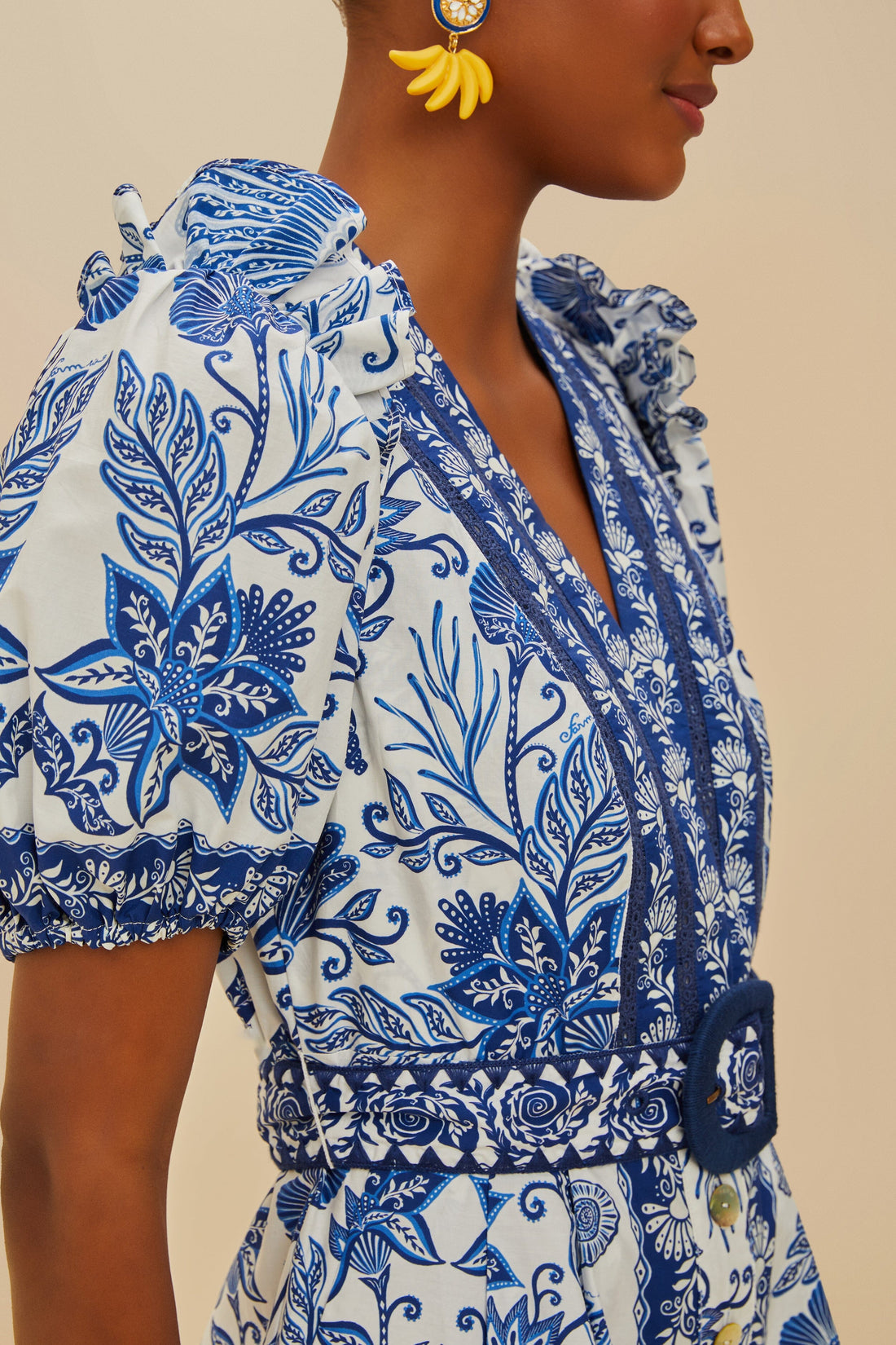 A woman wears the Farm Rio Shell Garden Off White Romper with blue florals, puff sleeves, a belted waist, and yellow floral earrings.