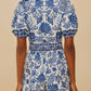 A person is seen from the back wearing the Farm Rio Shell Garden Off White Romper, featuring short sleeves and blue tropical florals, against a neutral background.
