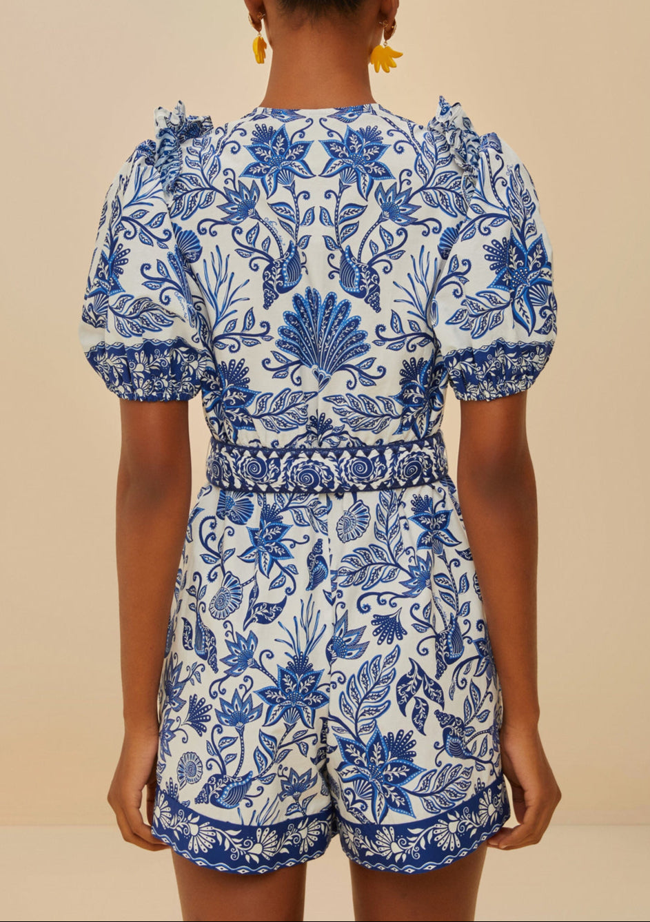 A person is seen from the back wearing the Farm Rio Shell Garden Off White Romper, featuring short sleeves and blue tropical florals, against a neutral background.