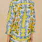 A woman is seen from behind wearing the Farm Rio Sicily Scarf Off White Shirt, a matching long-sleeve top and shorts set with bright yellow florals and blue scrolls on white, radiating tropical elegance.