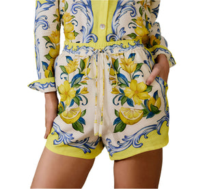 A person wears Farm Rio’s Sicily Scarf Off White Shorts, featuring a yellow lemon and floral print, an elastic drawstring waist, and hands in pockets.