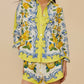 A woman wears Farm Rio's Sicily Scarf Off White Shorts, a matching set with a button-up shirt, featuring yellow floral and blue swirl patterns with lemons and leaves on a light background.