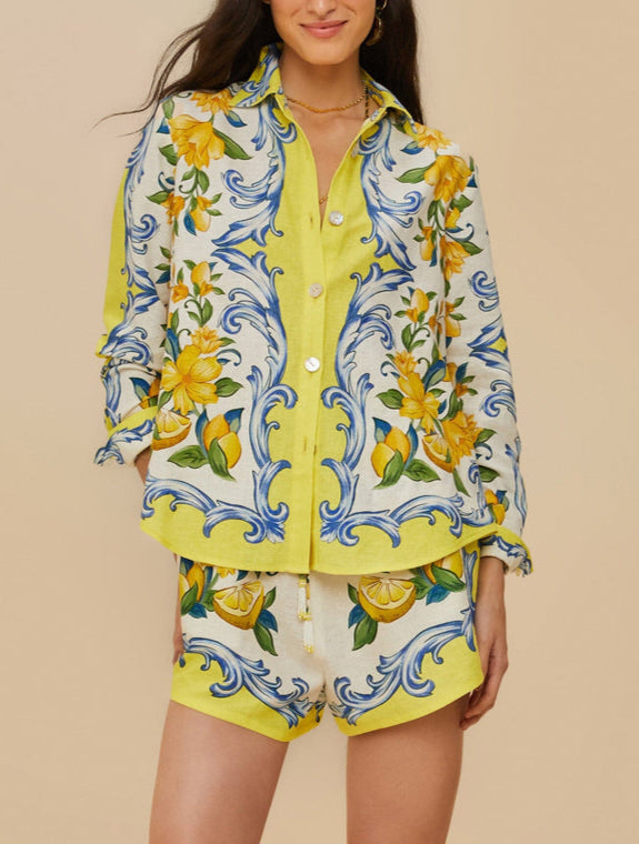 A woman wears Farm Rio's Sicily Scarf Off White Shorts, a matching set with a button-up shirt, featuring yellow floral and blue swirl patterns with lemons and leaves on a light background.