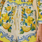 A person wears Farm Rio Sicily Scarf Off White Shorts featuring a yellow and blue floral and lemon print, elastic waistband, and drawstring tie. The matching long-sleeve shirt is partially visible.
