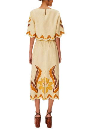 A woman is shown from the back wearing Farm Rio’s Summer Cutwork Sand Skirt, featuring short sleeves, yellow and brown floral appliqué, cutwork patterns, scalloped edges, and brown platform sandals.