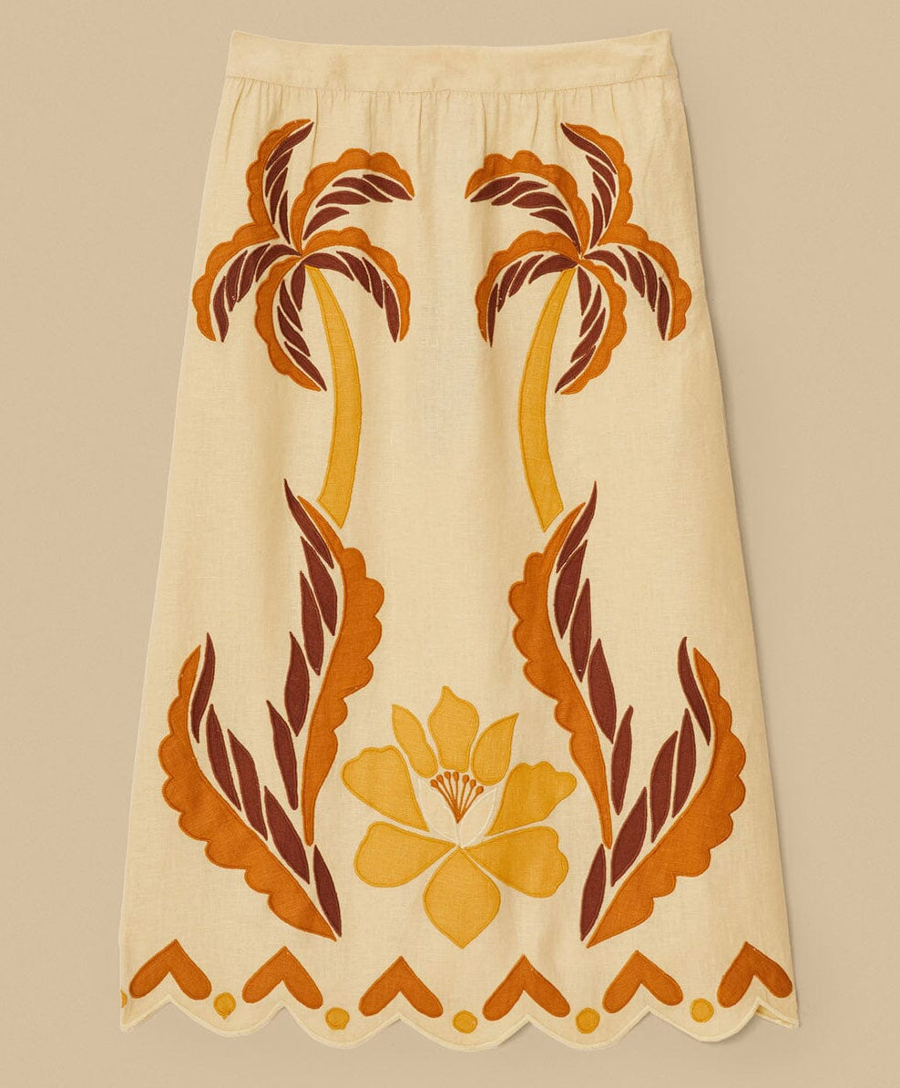 The Summer Cutwork Sand Skirt by Farm Rio features brown and yellow palm tree and floral appliqué with large leaves and a scalloped hem, inspired by FARM Rio’s signature cutwork patterns on a neutral beige background.