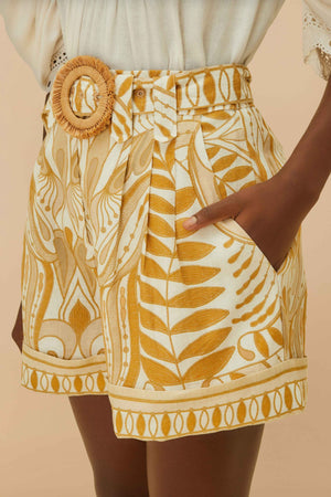 Wearing FARM Rio's Lace Nature Off-White Shorts, made from eco-responsible fibers, styled with a wide belt and a white blouse with lace sleeves that feature FARM Rio's iconic leafy patterns.