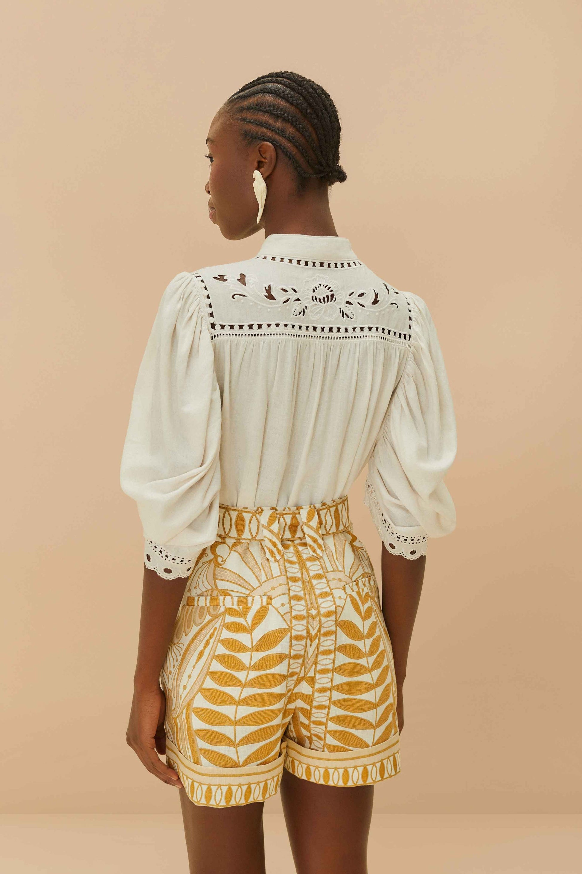 A person faces away, wearing a white lace-detailed blouse and the Lace Nature Off-White Shorts by Farm Rio, crafted from eco-responsible fibers, against a beige background.